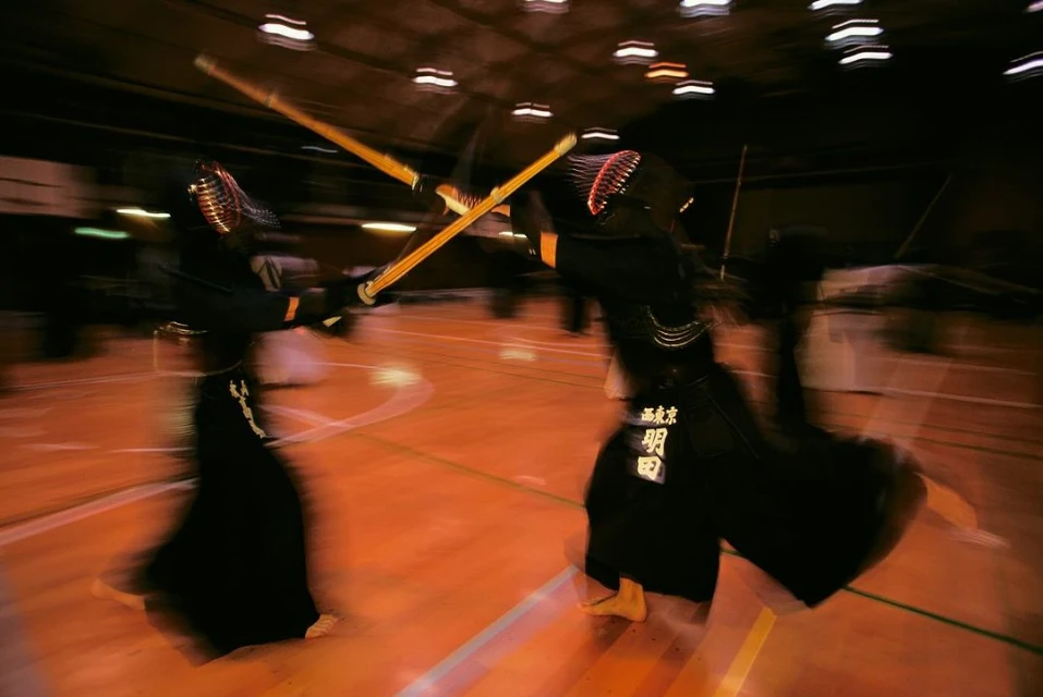 Two kendo fighters in protective gear compete in a dimly lit indoor space with blurred motion, wielding bamboo swords in a clash.