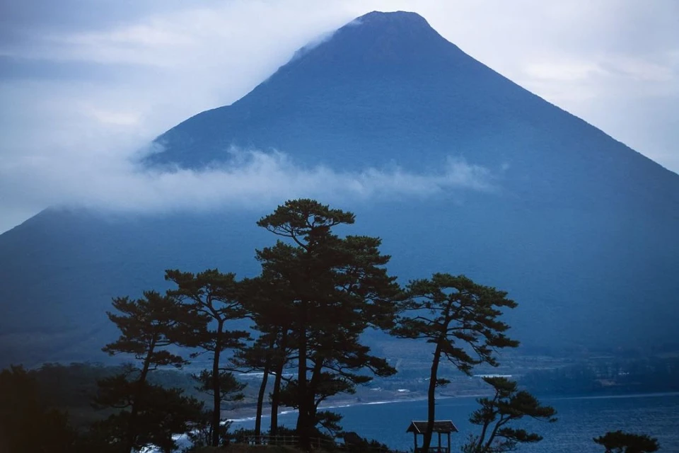 Scenic view of a large, blue-toned mountain partially obscured by low-lying clouds, framed by evergreen trees and a gazebo on a coast.