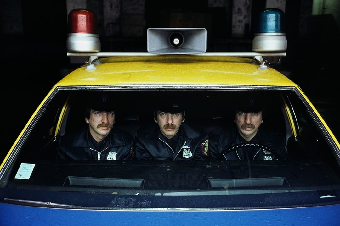Three adult males inside a yellow and blue police car with red and blue lights and a loudspeaker on top, wearing black hats and uniforms.