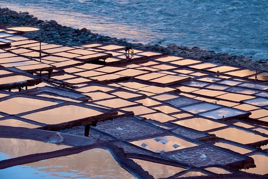 Scenic view of terraced salt ponds with workers near a river during the day, reflecting light and shadows on the water and land.