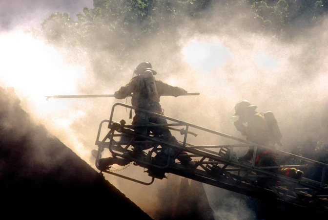Two firefighters stand on a ladder amidst smoke and water, battling a fire, demonstrating fire safety and emergency response.