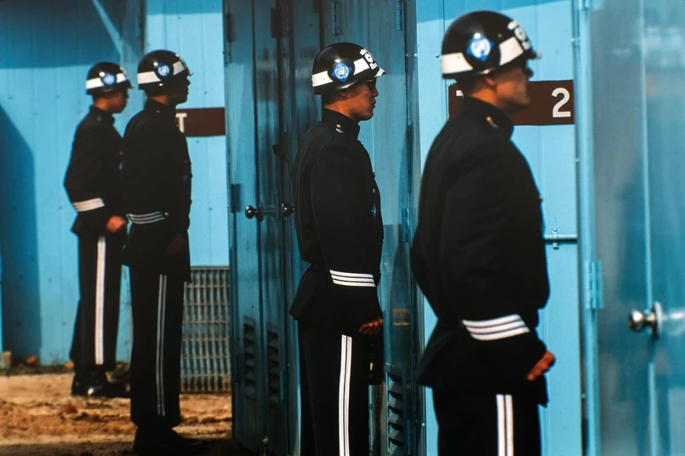 Four guards in dark uniforms and helmets stand at attention in front of light-blue building with doors in an outdoor setting.
