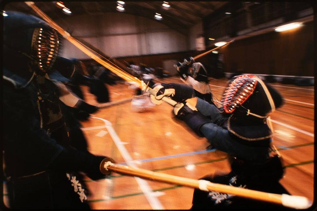 Kendo martial artists in protective gear compete with bamboo swords on a polished wooden floor in a brightly lit indoor gymnasium.