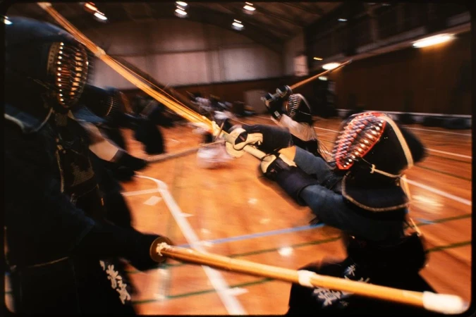 Kendo martial artists in protective gear compete with bamboo swords on a polished wooden floor in a brightly lit indoor gymnasium.