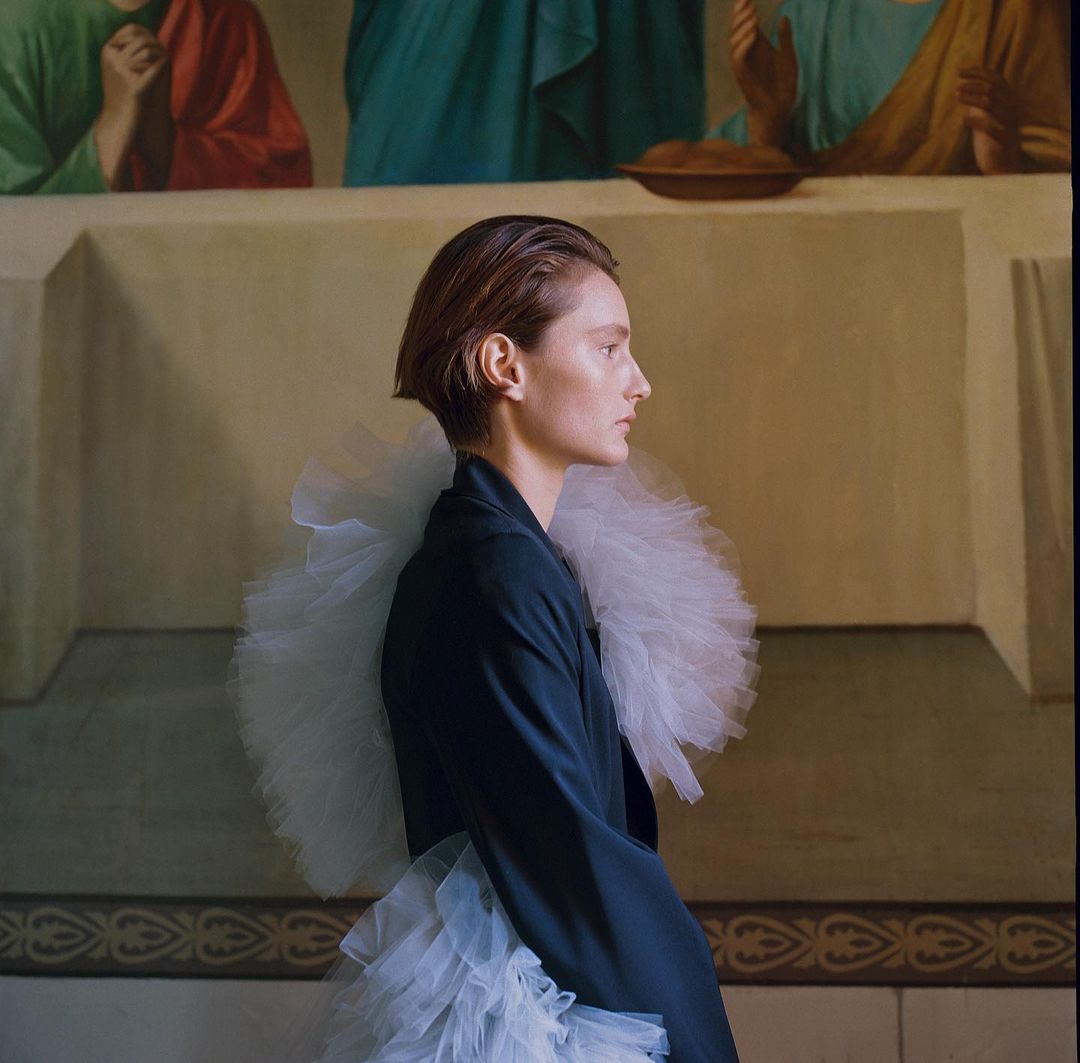 Profile shot of a female model wearing a black blazer and white tulle collar and skirt against a backdrop of a religious mural.