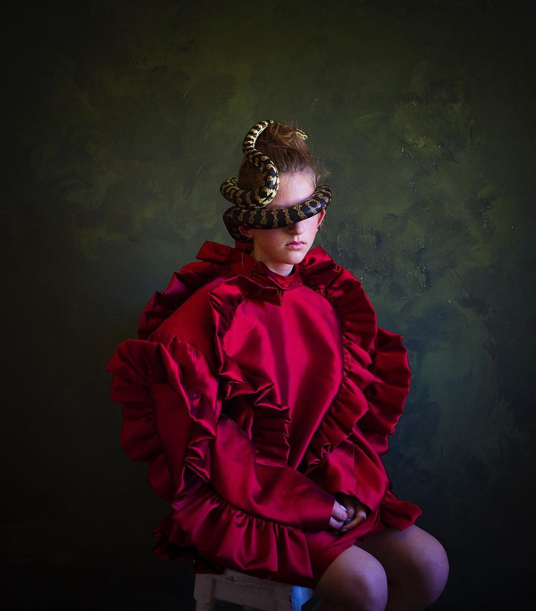 Portrait of a girl with a snake wrapped around her head, wearing a red ruffled dress against a dark background in a studio.