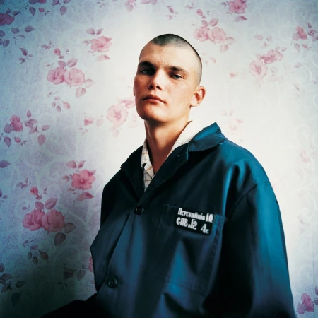 Portrait of a young man with a shaved head, wearing a dark blue jacket, standing indoors against a floral patterned wallpaper background.