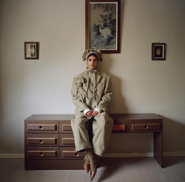 A person wearing an oversized beige suit and hat sits on a wooden dresser, posing in front of a wall with vintage artwork.