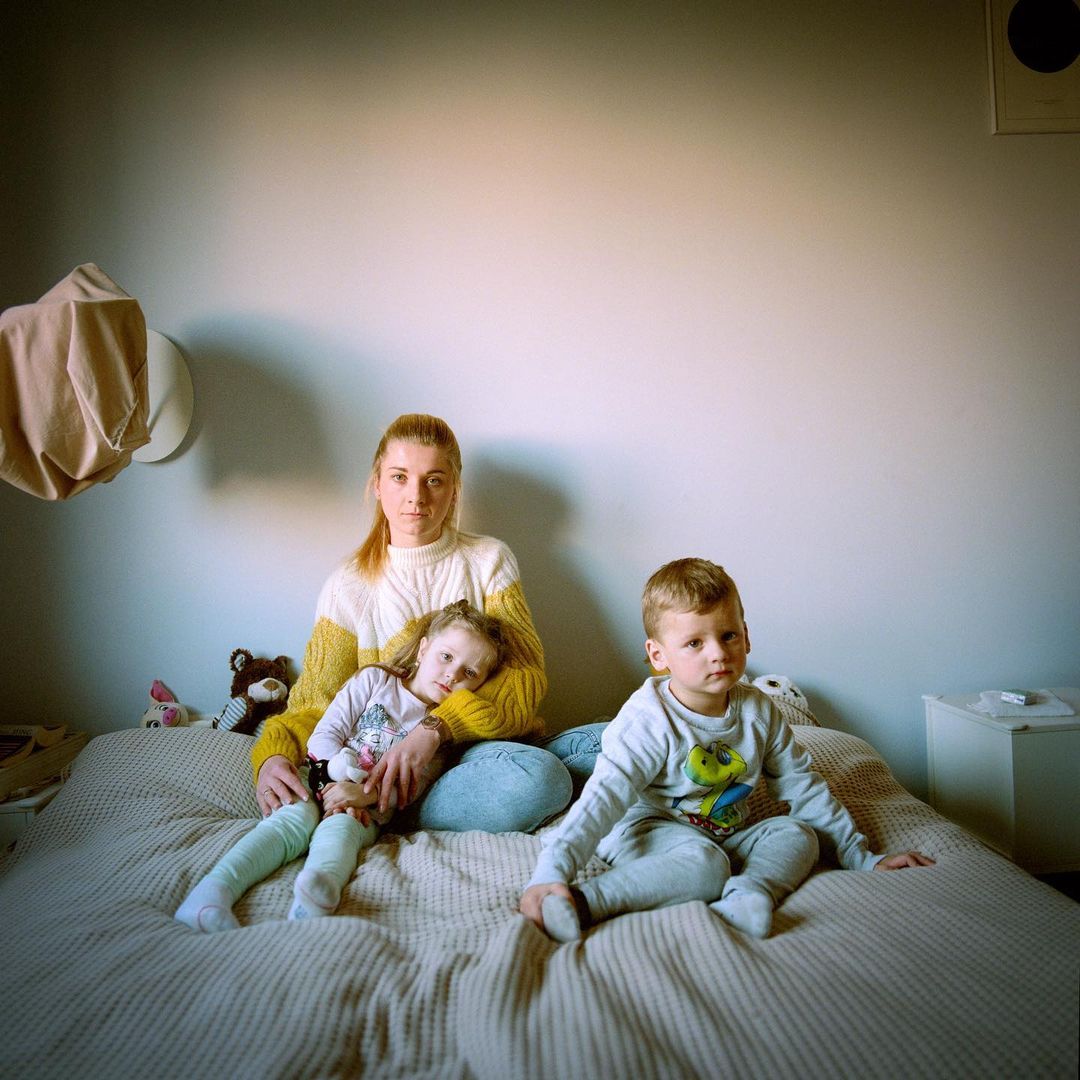 Indoor portrait of a mother sitting on a bed with her two young children, featuring neutral tones and soft lighting in a home setting.