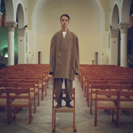 Young adult in an oversized brown suit standing on a chair in a church, looking at camera with rows of empty wooden chairs in the background.