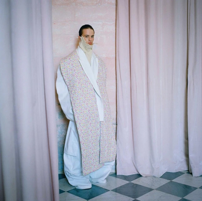 A person poses indoors wearing a white and floral print jacket, a white turtleneck, and white pants against a patterned tile floor.