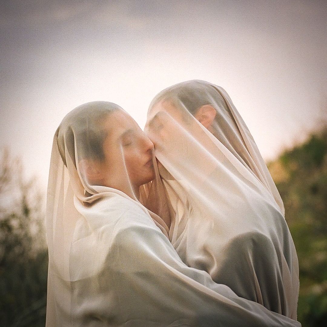 Close up portrait of two adults kissing under a sheer fabric, soft focus, and natural lighting with greenery visible in the background.