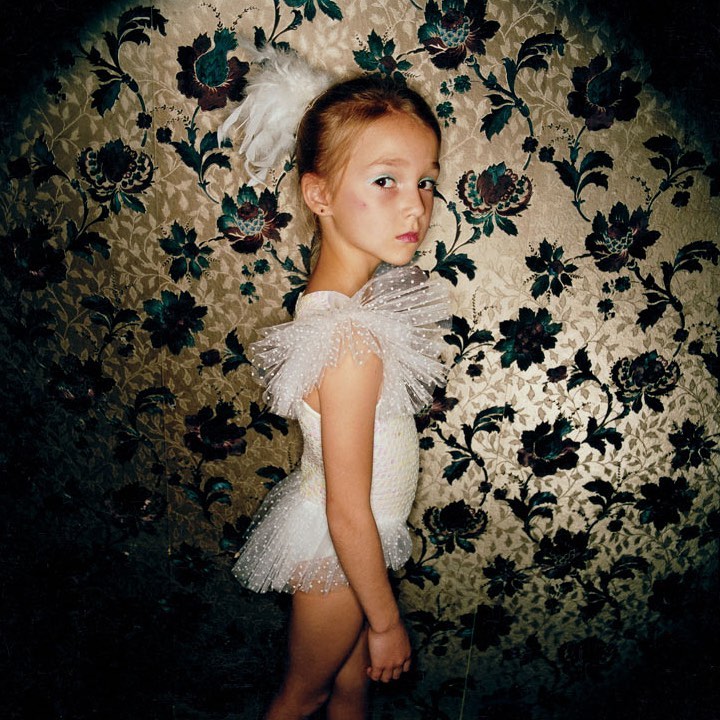 Young girl in white leotard stands against a floral patterned wall with a somber expression in a dimly lit indoor setting.