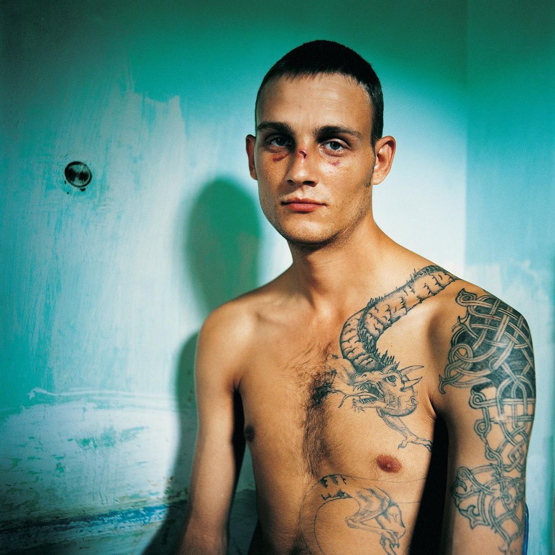 Striking portrait of a bare-chested man with visible facial injuries and prominent tattoos in a minimally decorated room, intense gaze.