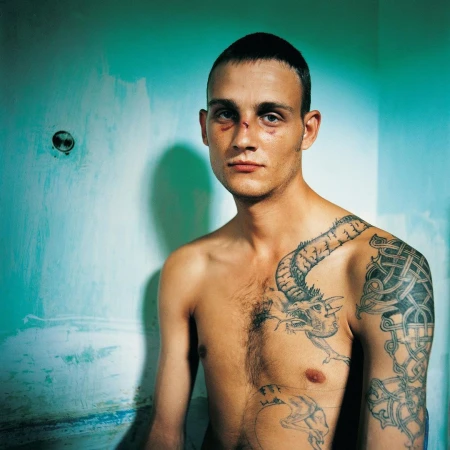Striking portrait of a bare-chested man with visible facial injuries and prominent tattoos in a minimally decorated room, intense gaze.