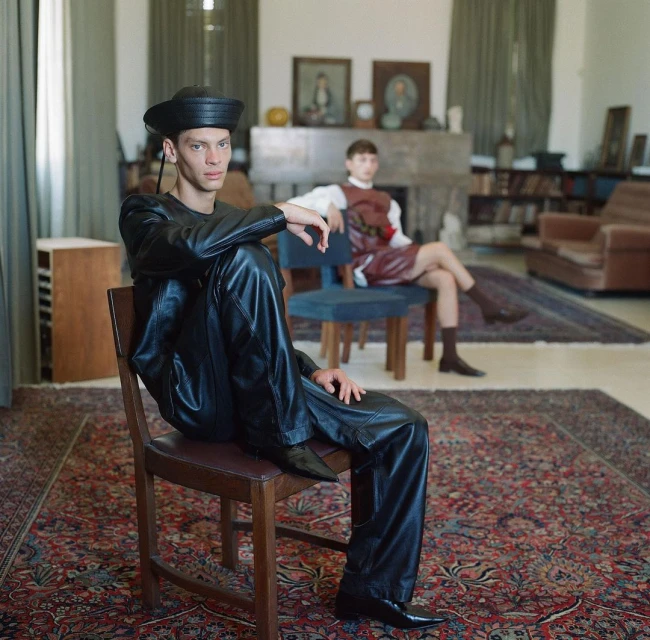 Two young men in vintage fashion posing indoors. One man in black leather sits on a chair; the other man sits in the background.