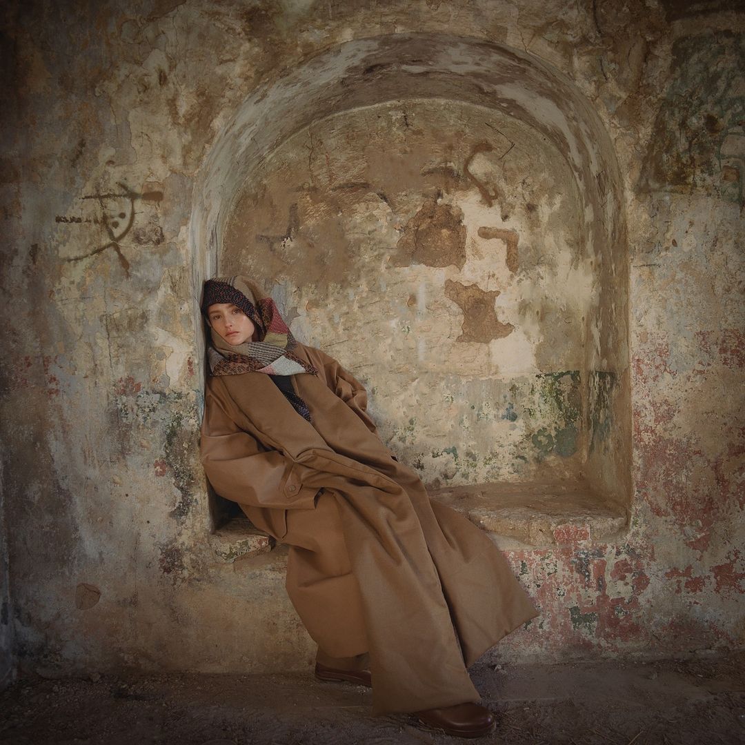 Woman sits in a stone archway wearing a long brown coat, patterned scarf, and hat, with neutral tones and soft lighting.