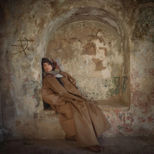 Woman sits in a stone archway wearing a long brown coat, patterned scarf, and hat, with neutral tones and soft lighting.