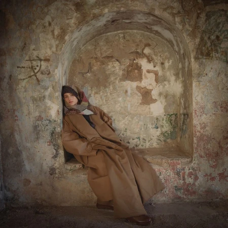 Woman sits in a stone archway wearing a long brown coat, patterned scarf, and hat, with neutral tones and soft lighting.