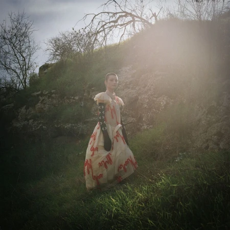 Woman in vintage dress poses in natural light among grassy hills. The scene is bathed in sunlight with a touch of post-production.