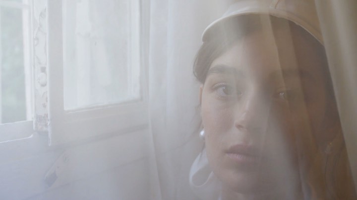 Close-up of a woman's face partially obscured by a sheer white curtain next to a window, with soft, diffused lighting.