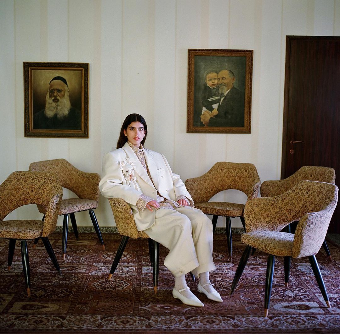 A young adult woman in a cream-colored pantsuit sits on a patterned chair surrounded by similar chairs in a room with paintings.