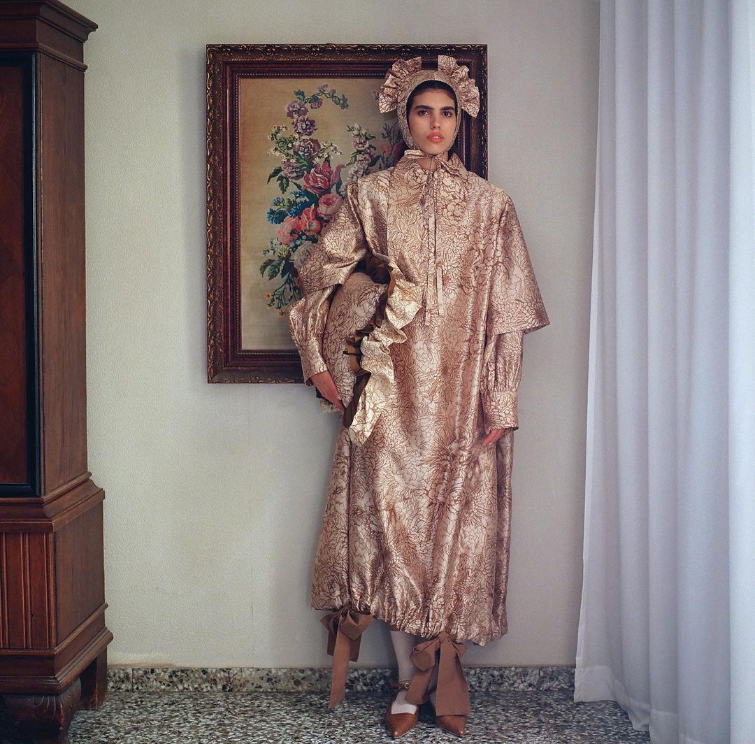 Woman in an ornate, patterned gold gown with ruffled bonnet stands in an interior with vintage floral painting and dark wood cabinet.