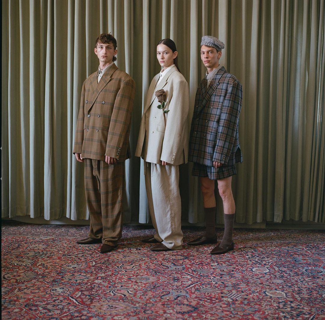 Three people wearing oversized plaid suits stand on a patterned carpet in front of a muted green curtain in a studio fashion shot.