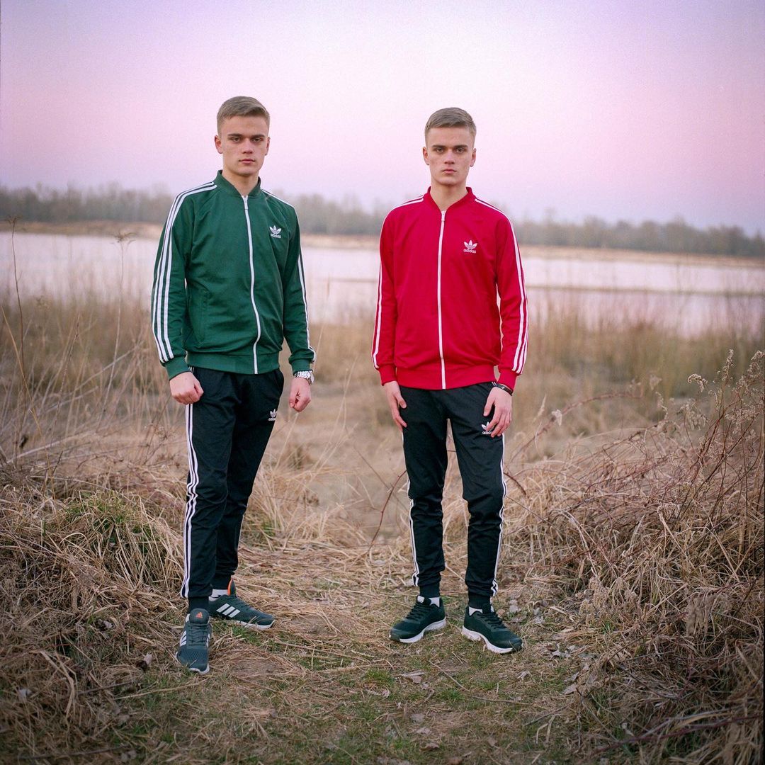 Two young men stand side-by-side outdoors, wearing matching sporty tracksuits. Tall dry grass is around them and a lake in the background.