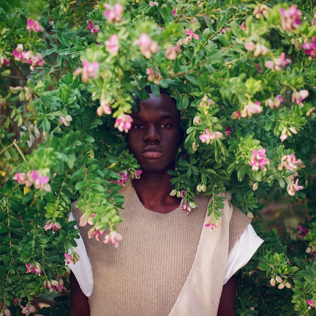 Portrait of a Black man surrounded by flowering green bushes with pink blossoms in natural light, wearing a sleeveless top.