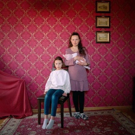 Young girl seated on a chair beside a woman holding a baby in a room with patterned pink damask wallpaper and a patterned rug.