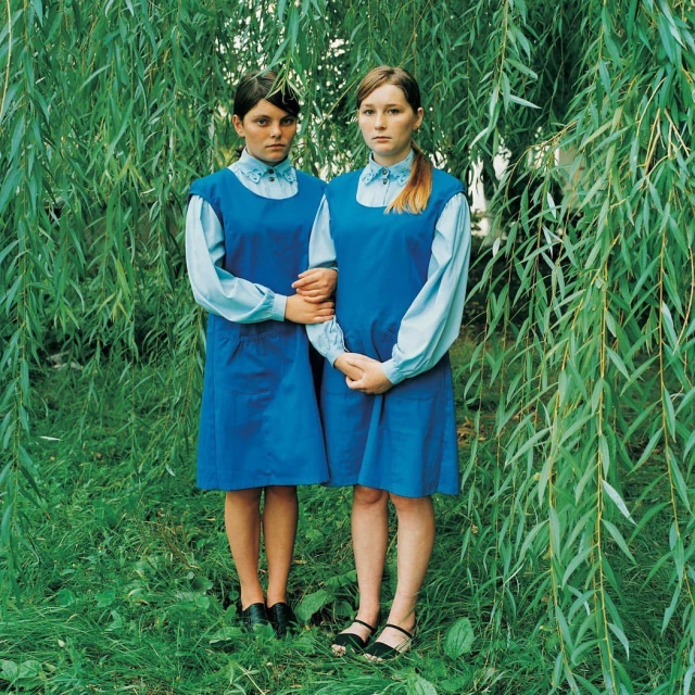 Two fair-skinned young women in blue dresses stand in front of a willow tree in a natural setting, wearing similar vintage clothing.