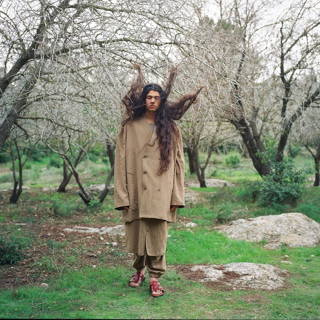 A person stands in an autumnal forest landscape, wearing a brown coat, matching pants, and red sandals, with hair extending to the sides.