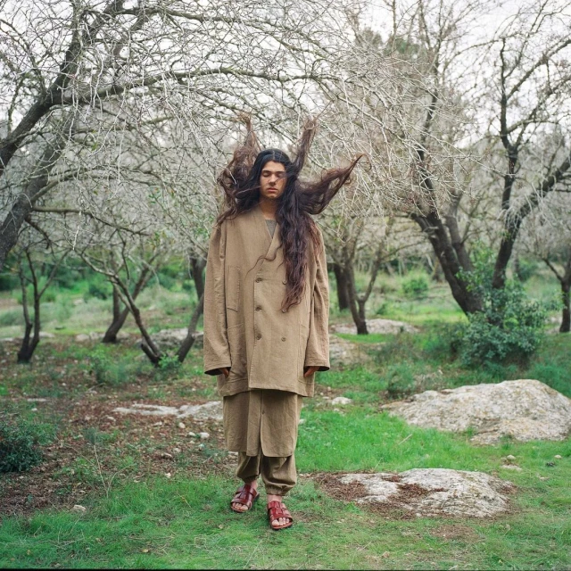A person stands in an autumnal forest landscape, wearing a brown coat, matching pants, and red sandals, with hair extending to the sides.