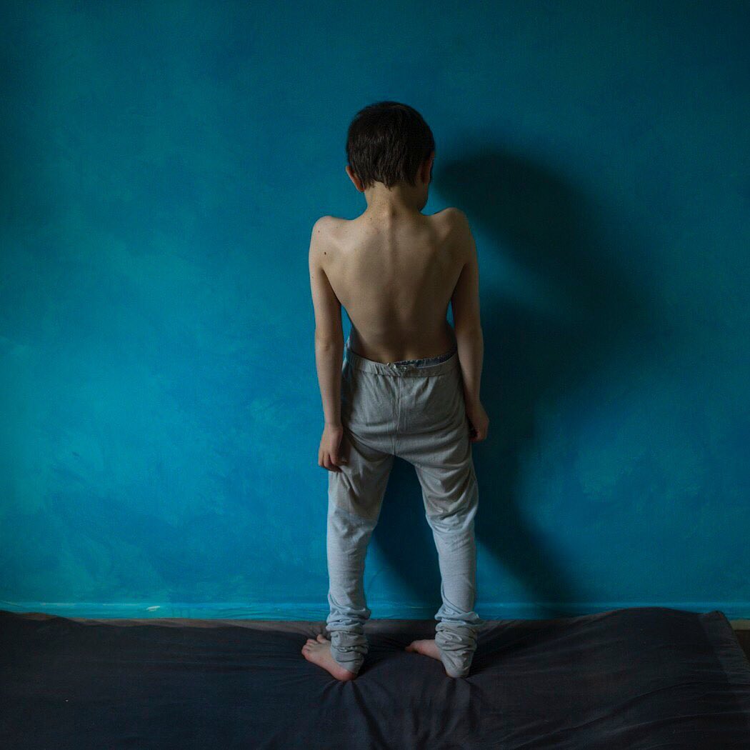 Young boy stands facing a textured blue wall, shot from the back, wearing gray sweatpants on a gray mat in an indoor setting.