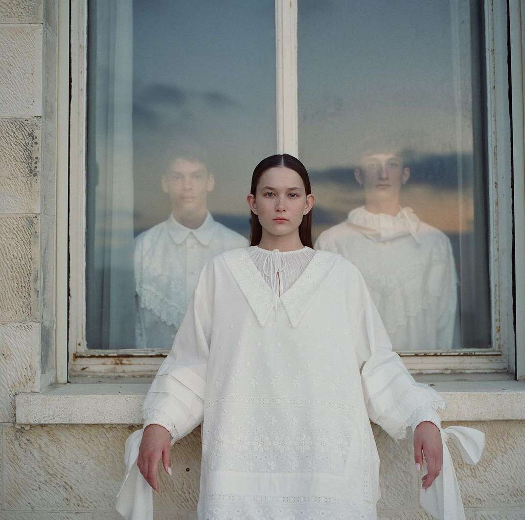 Indoor fashion portrait of a young woman in an embroidered white dress, with two blurred figures reflected in the window behind her.