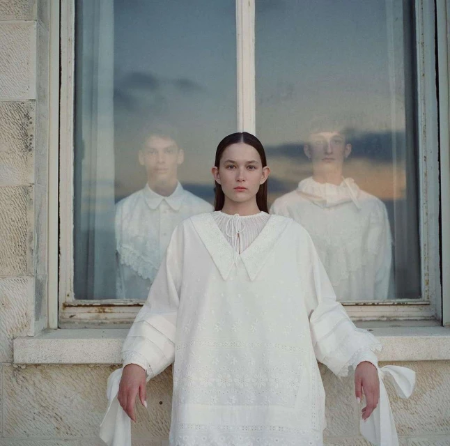 Indoor fashion portrait of a young woman in an embroidered white dress, with two blurred figures reflected in the window behind her.
