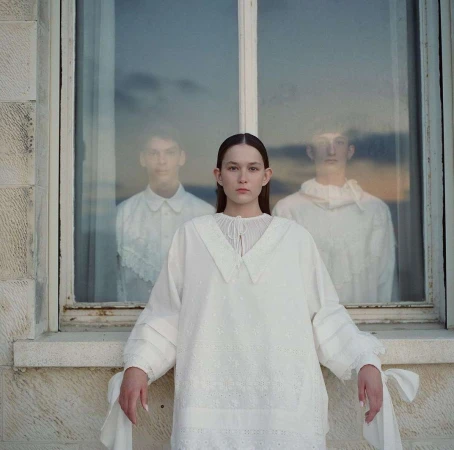 Indoor fashion portrait of a young woman in an embroidered white dress, with two blurred figures reflected in the window behind her.