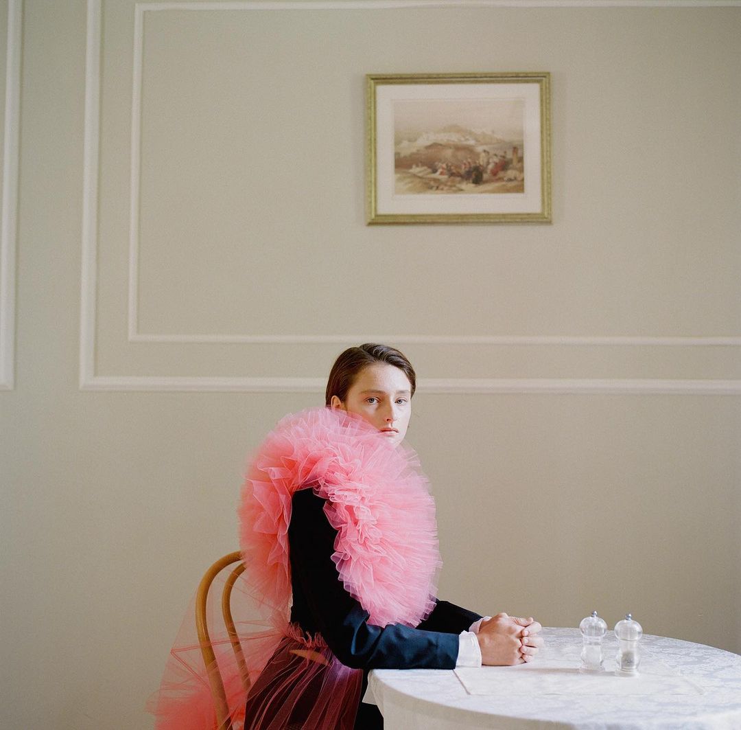 A woman in a dark suit and pink tulle collar and skirt sits at a table in a dining room with a landscape painting on the wall.