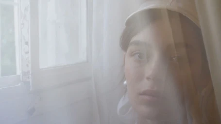 Portrait of a woman wearing a white cap looking towards an open window with soft hazy light filtering through curtains.