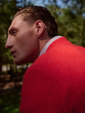 Close-up profile of a man with slicked hair wearing a bright red wool sweater and a small silver hoop earring outdoors.