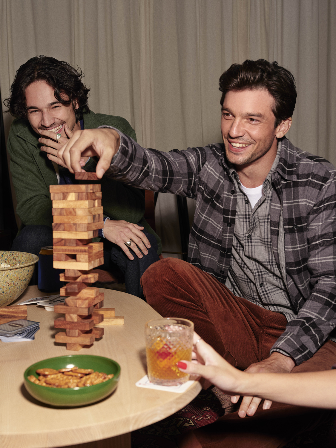 Two men playing a wooden block tower game indoors, one man reaching to move a piece, while another person holds a cocktail glass nearby.