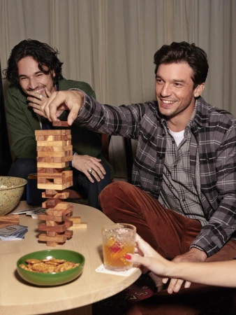 Two men playing a wooden block tower game indoors, one man reaching to move a piece, while another person holds a cocktail glass nearby.