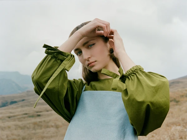 Portrait of a woman with long hair adjusting her ponytail, wearing a green blouse with tied cuffs and a light blue pinafore outdoors.