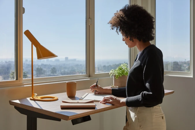 A woman with curly dark hair stands at a wooden standing desk by a large window, writing in a notebook next to a yellow lamp.