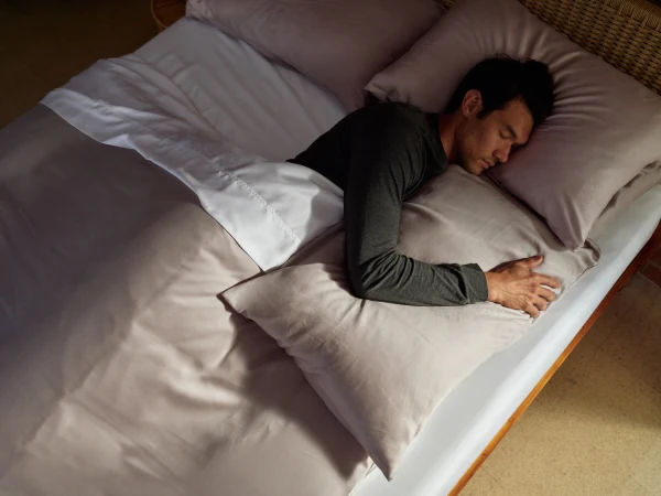 A man wearing a long-sleeved dark gray shirt sleeps peacefully in bed, his arm resting on a light taupe pillow under a white sheet.