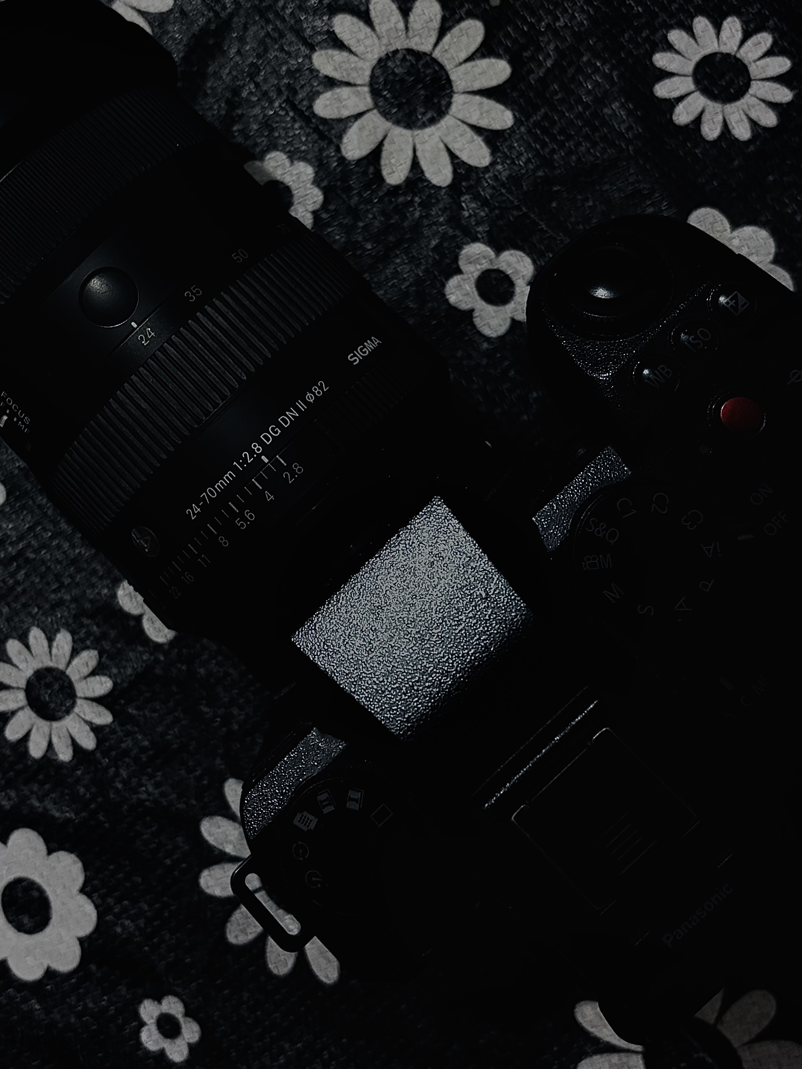 Close-up of a black camera and lens resting on a dark patterned fabric background with white flower motifs.
