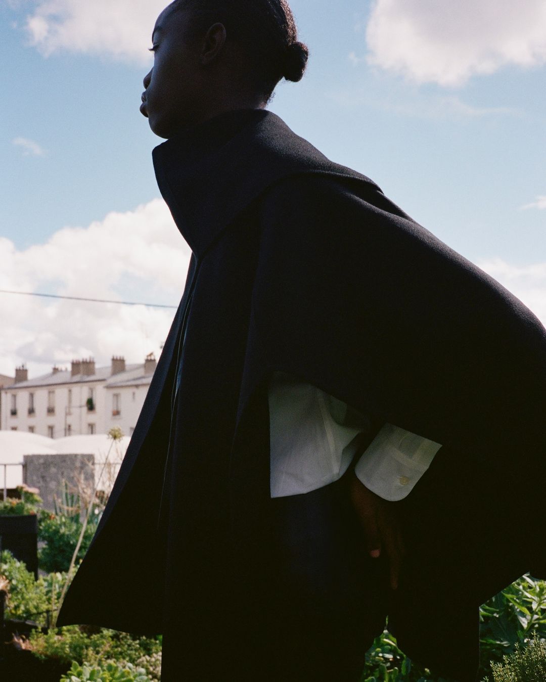 An African American female model in a dark coat stands in a garden with apartment buildings and a cloudy blue sky in the background.