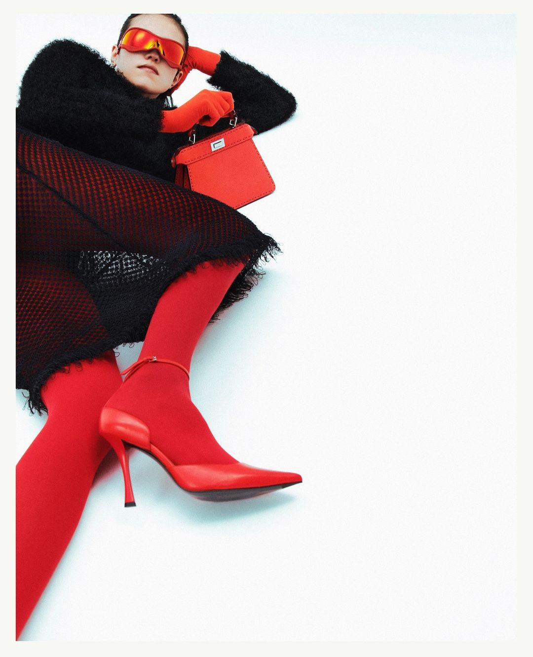 Adult female fashion model in a black top, red tights and shoes holding a red purse, laying down in a light studio setting.