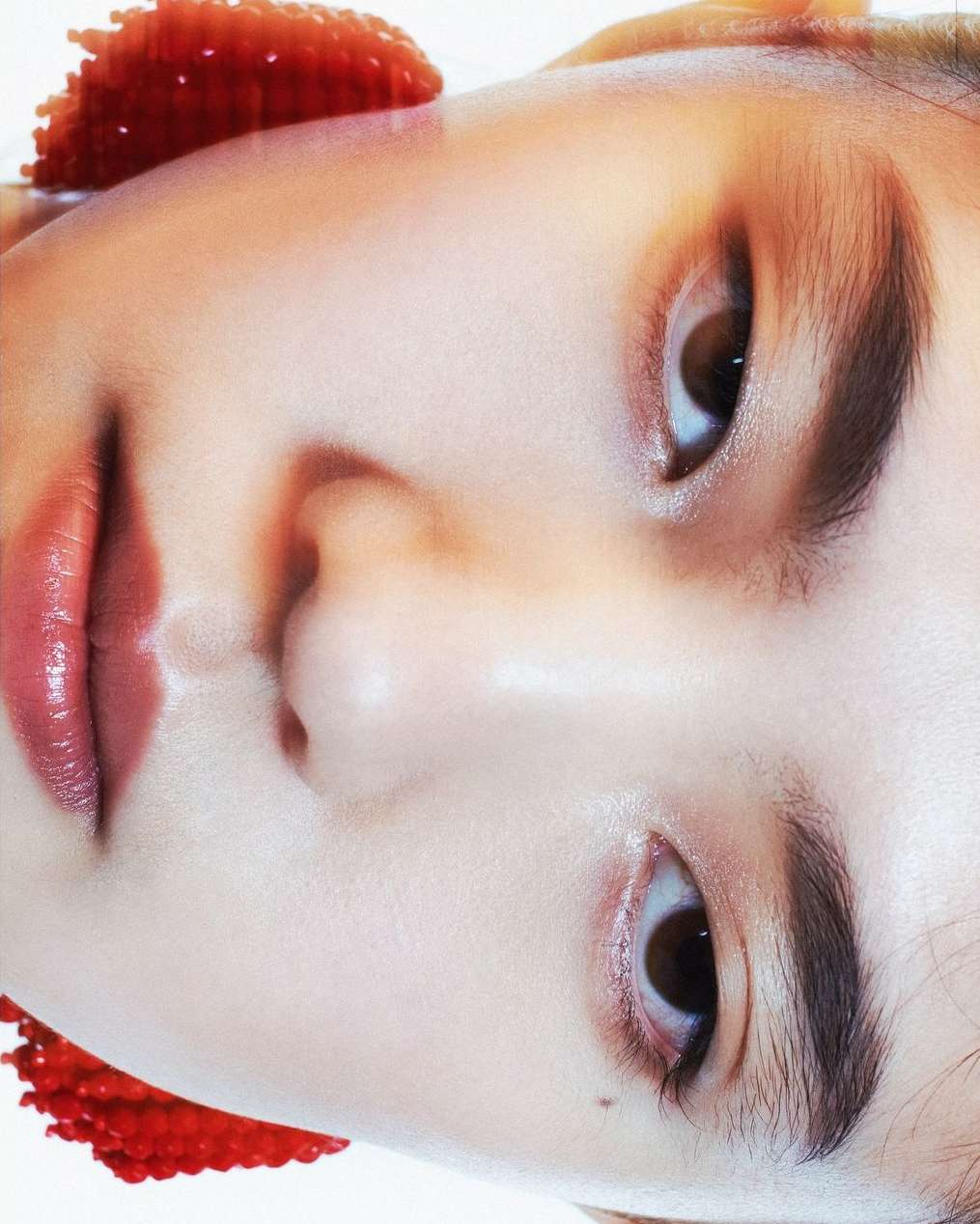 Close-up of a young adult woman's face with smooth skin, pink lips, and soft lighting, with red beaded details on the edges.